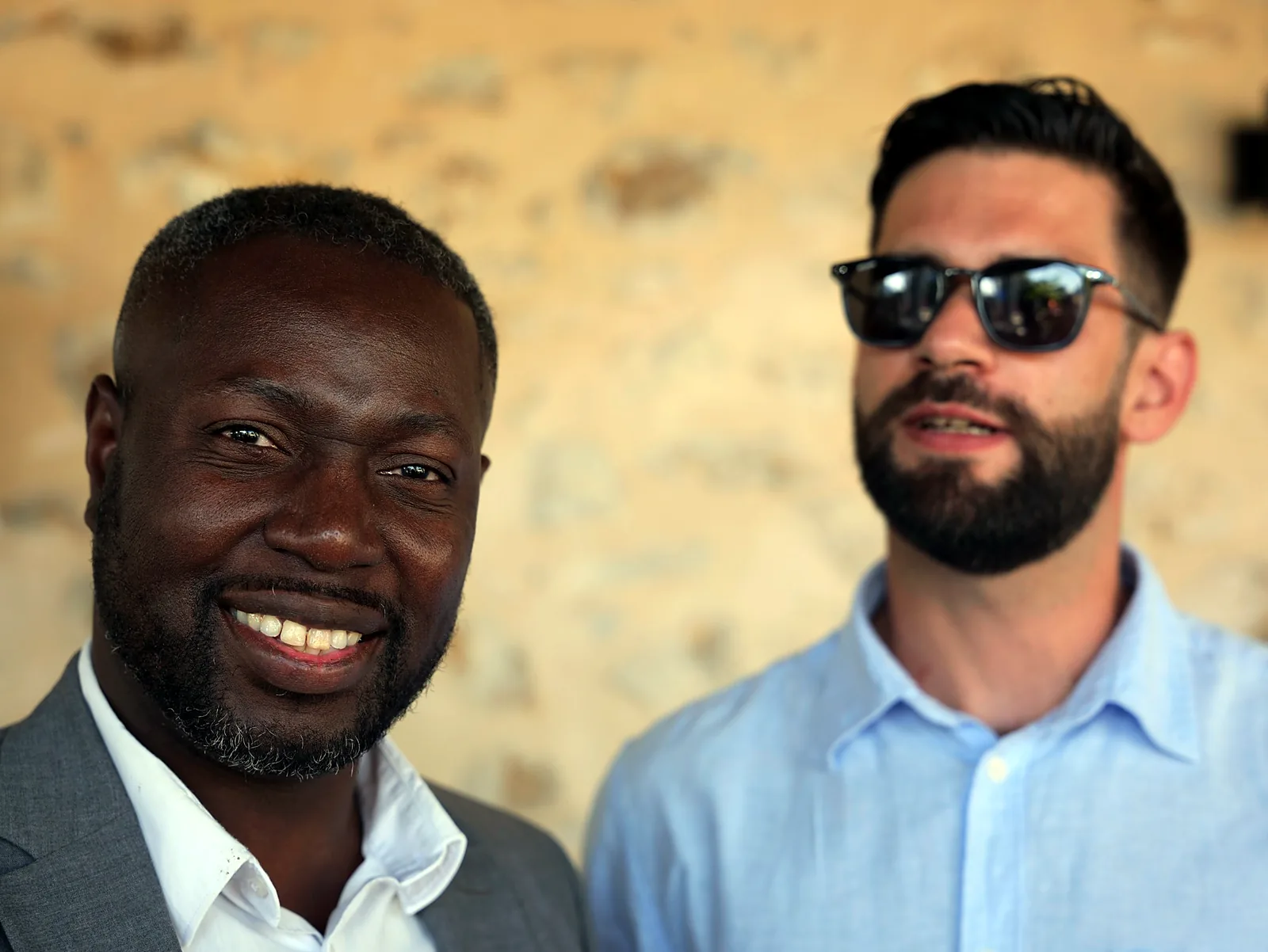 Deux hommes posent côte à côte et sourient lors d'un événement, devant un mur de pierre